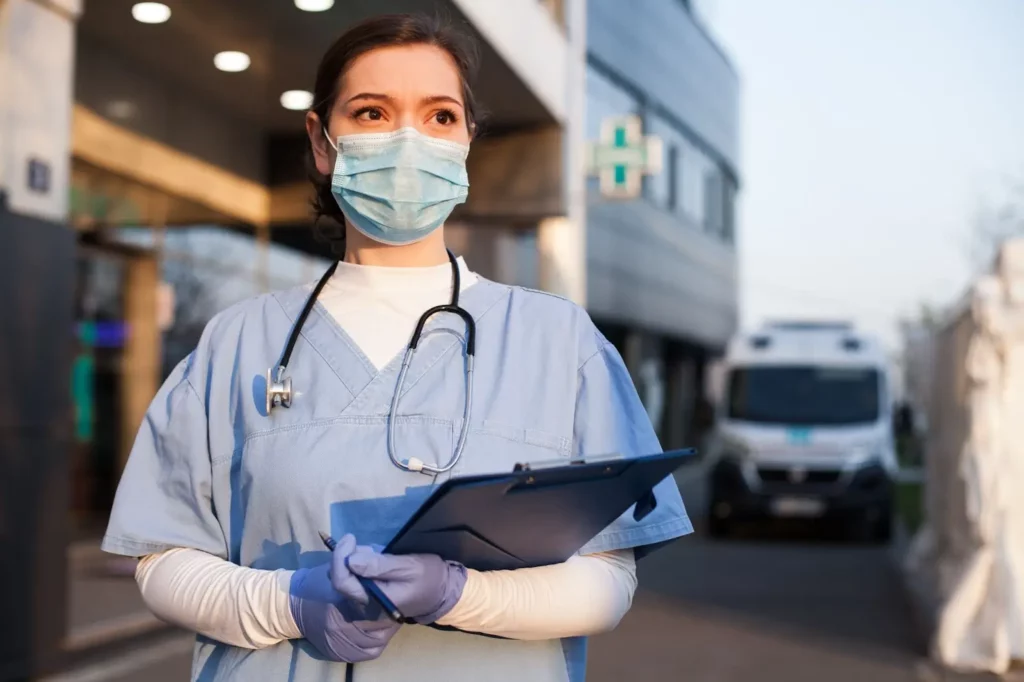 woman doctor with face mask on holding a clipboard infront of hospital